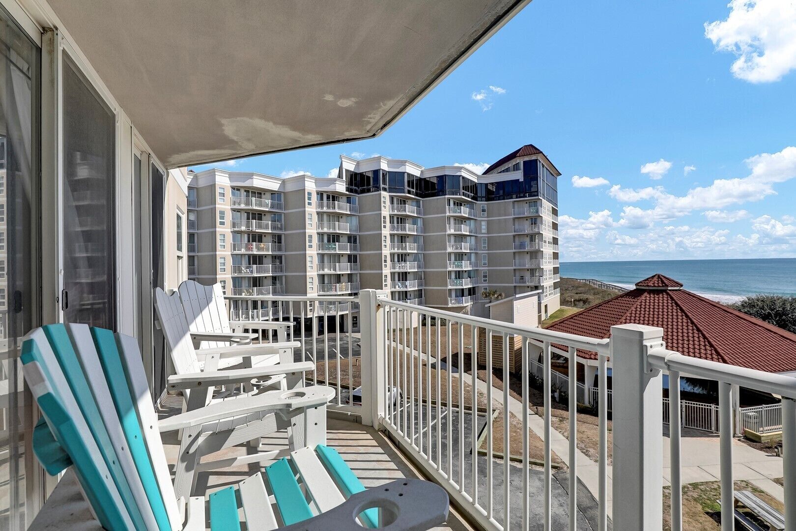 Ein Balkon mit weißen Geländern und zwei gestreiften Stühlen, mit Blick auf ein Gebäude am Strand, das mehrere Balkone und ein rot gedecktes Gebäude aufweist.