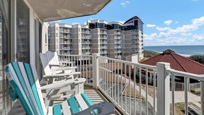 Een balkon met witte reling en twee gestreepte stoelen, met uitzicht op een gebouw aan het strand met meerdere balkons en een structuur met een rood dak.