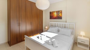 Un dormitorio con un armario de madera, una cama con ropa de cama blanca, una mesita de noche con una lámpara y un cuadro en la pared.