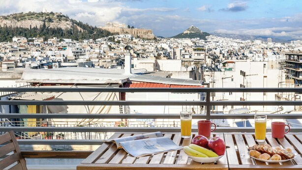Una mesa con alimentos para el desayuno y un periódico en un balcón con vistas a un paisaje urbano con edificios históricos.