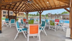 Une terrasse extérieure avec des chaises blanches et colorées, un ventilateur de plafond et une vue sur une piscine.