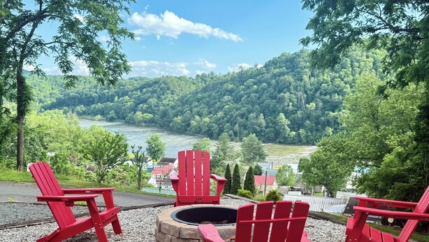A seating area with red Adirondack chairs and a stone fire pit, overlooking a river and forested hills. - If you”re looking for that perfect weekend get away, you just found it! (Fries)