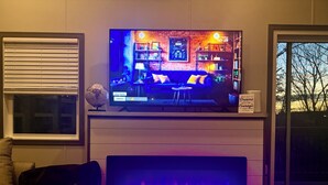 A living room with a flat-screen TV mounted on a wall, a fireplace below it, and a window to the right. - If you”re looking for that perfect weekend get away, you just found it! (Fries)
