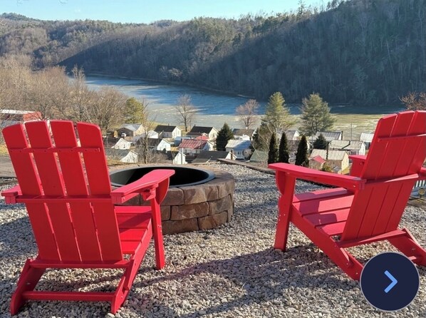 Zwei rote Adirondack-Stühle, die einem Feuerkorb mit Blick auf einen See und bewaldete Hügel zugewandt sind.