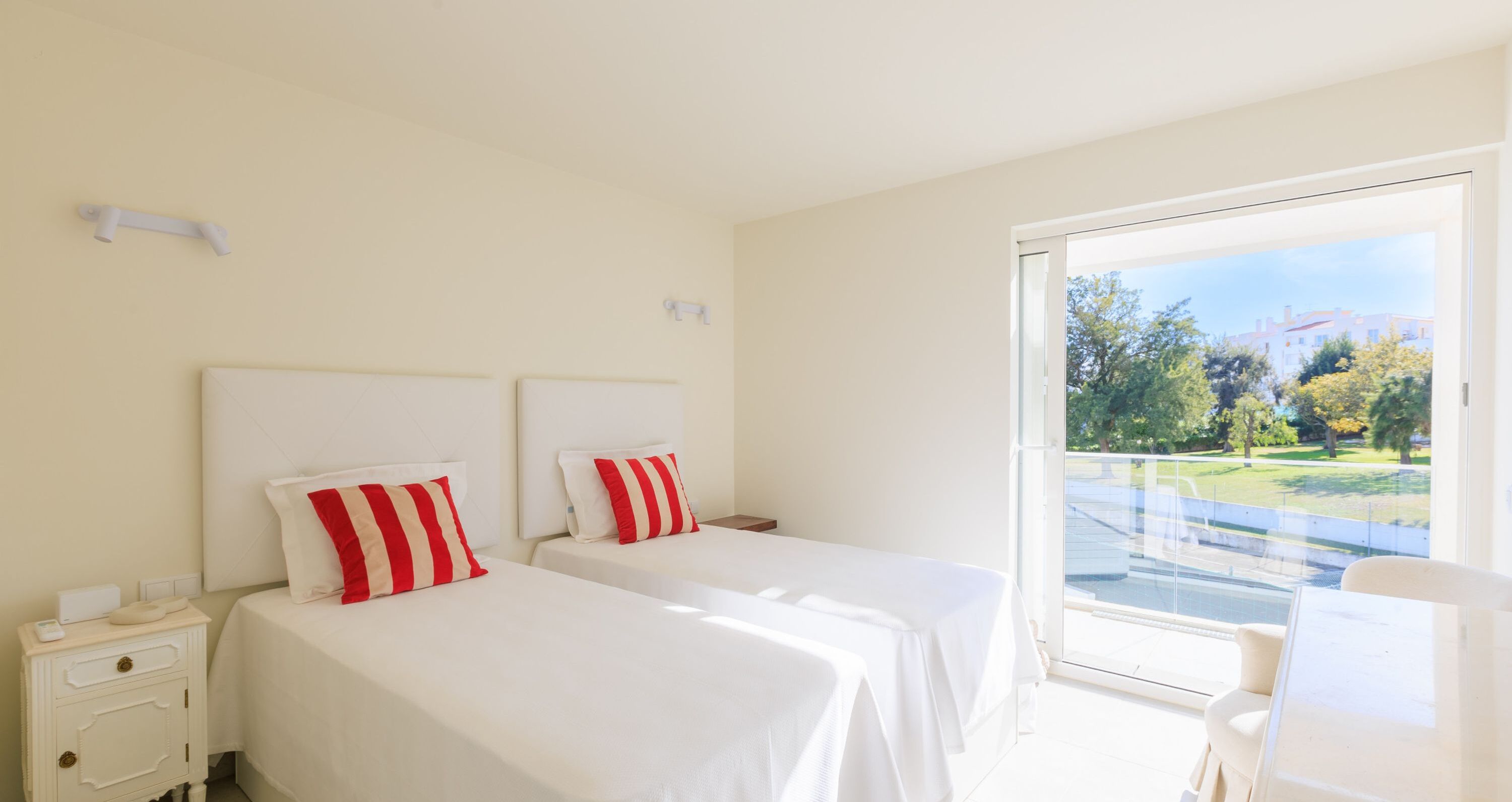 A hotel room with two beds, a small desk, and a balcony with a view of a park and buildings.
