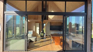 Ein modernes Interieur mit einer großen Schiebeglasdoor, die auf eine Holzterrasse führt. Innenbereich mit Küchenzeile, Esstisch und Stühlen sowie einer Sitzecke mit Sofa und Kissen.