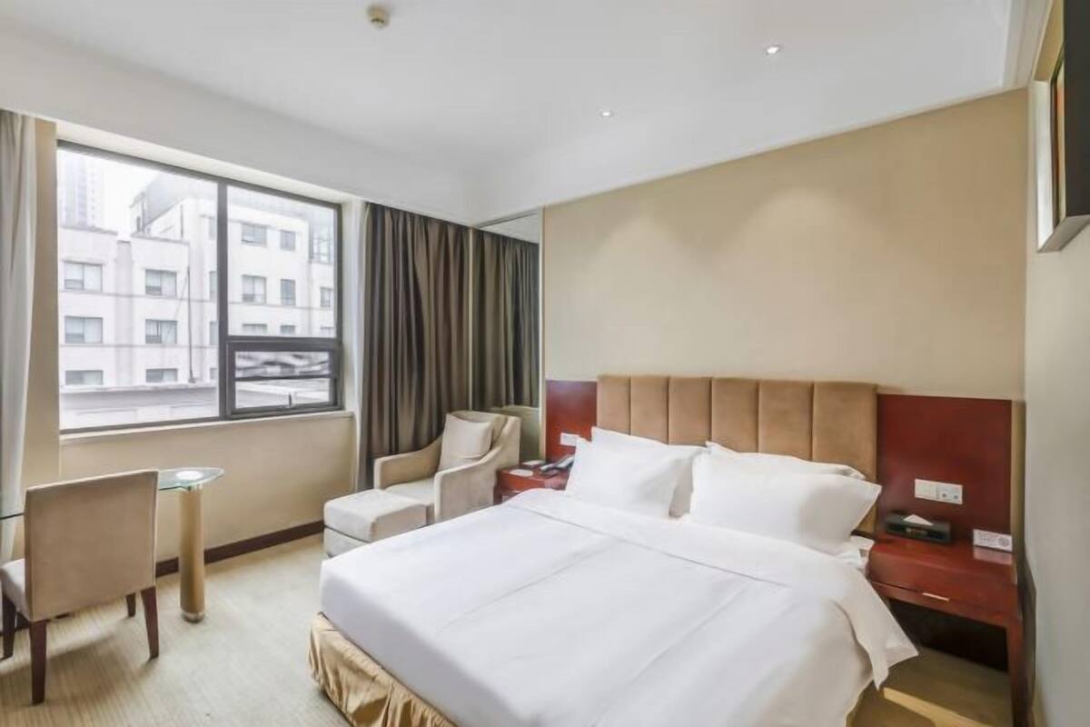 Quarto de hotel com cama, escrivaninha, cadeira, mesa de cabeceira e janela com cortinas.