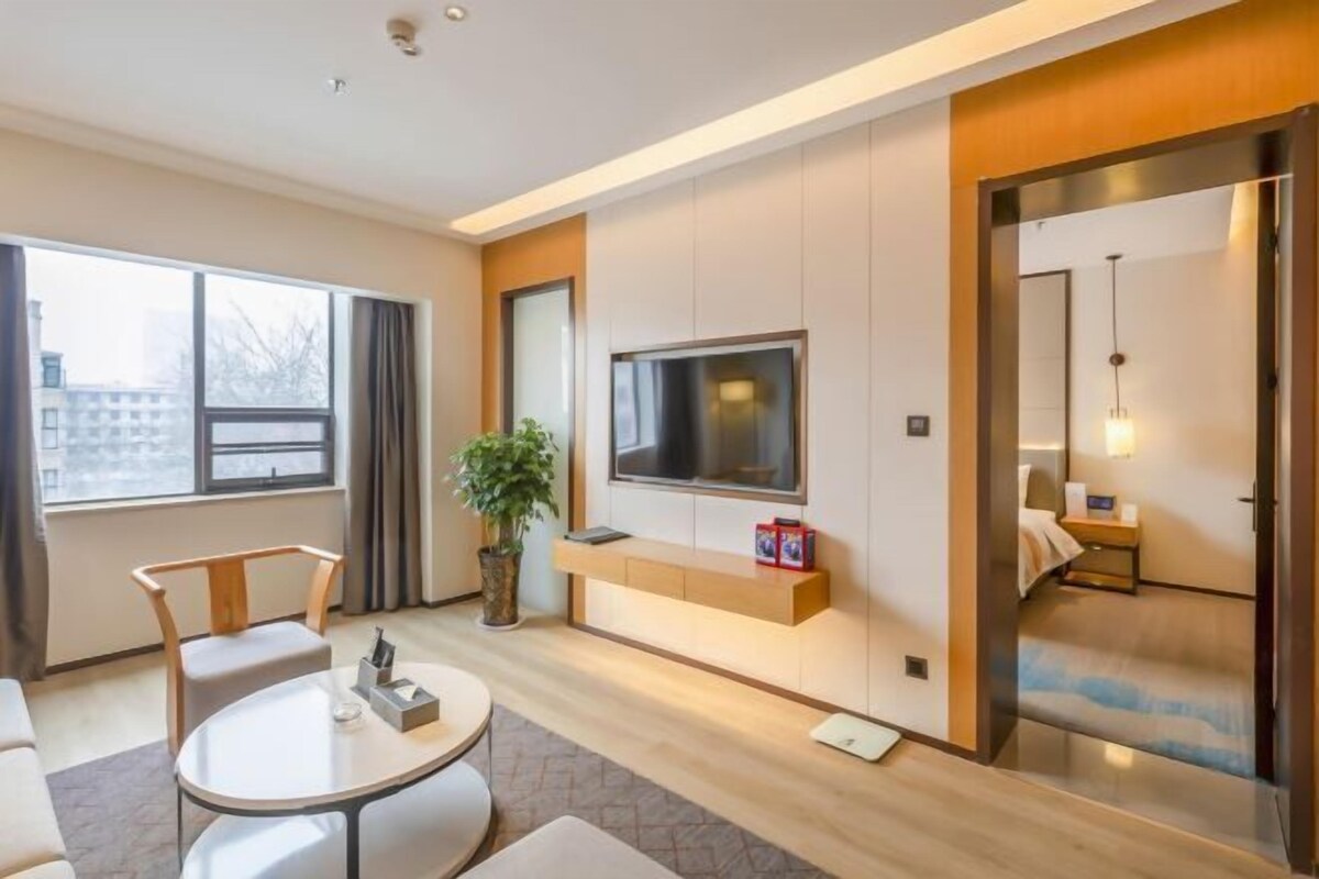 Una habitación de hotel moderna con un ventanal grande, un televisor de pantalla plana montado en la pared, un rincón de descanso con una mesa redonda y una planta pequeña.