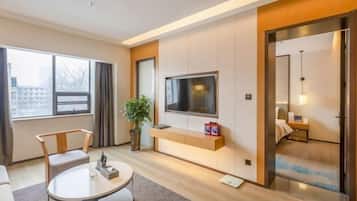 A modern hotel room with a large window, a flat-screen TV mounted on the wall, a seating area with a round table, and a small plant.