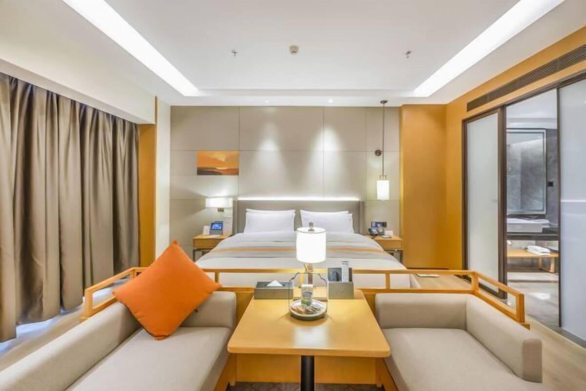 Quarto de hotel com cama, mesa com abajur, sofá com almofada laranja e uma janela grande.