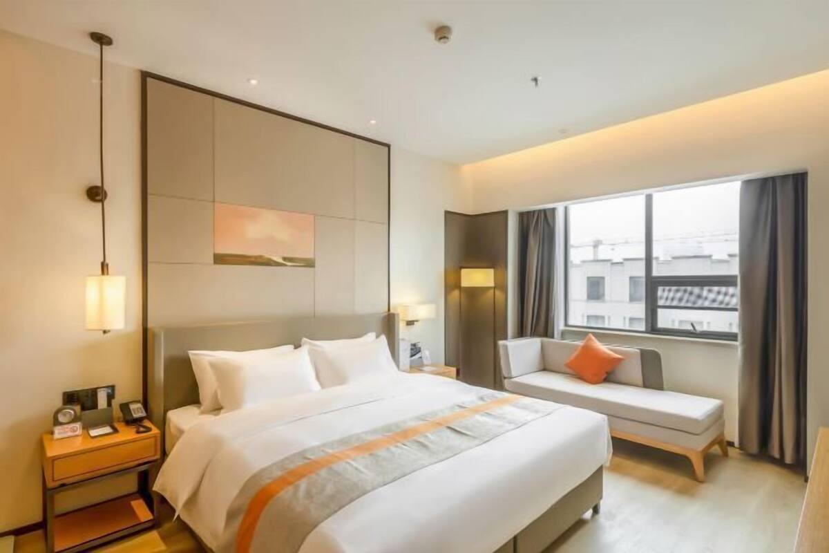 Quarto de hotel com uma cama, mesa de cabeceira, poltrona, sofá, janela e uma obra de arte pendurada na parede.