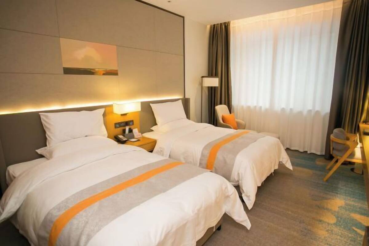 Quarto de hotel com duas camas, uma mesa, uma cadeira, uma luminária e uma janela com cortinas.
