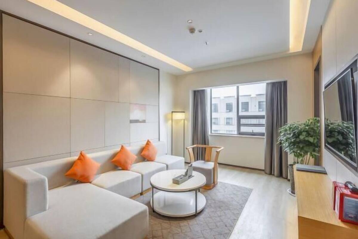 Quarto de hotel moderno com sofá, mesa de centro redonda, escrivaninha e uma janela grande com cortinas.