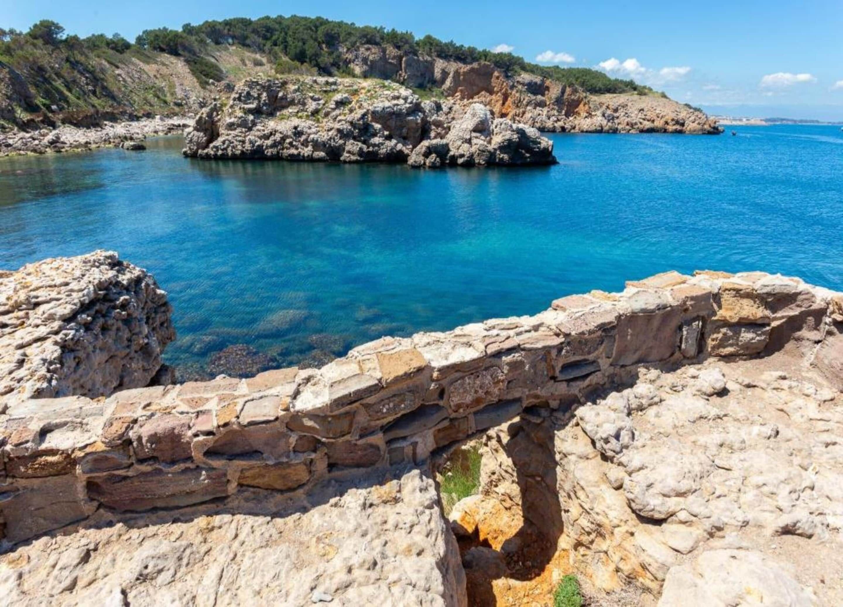 A coastal stone structure with a clear blue sea and rocky terrain in the background.