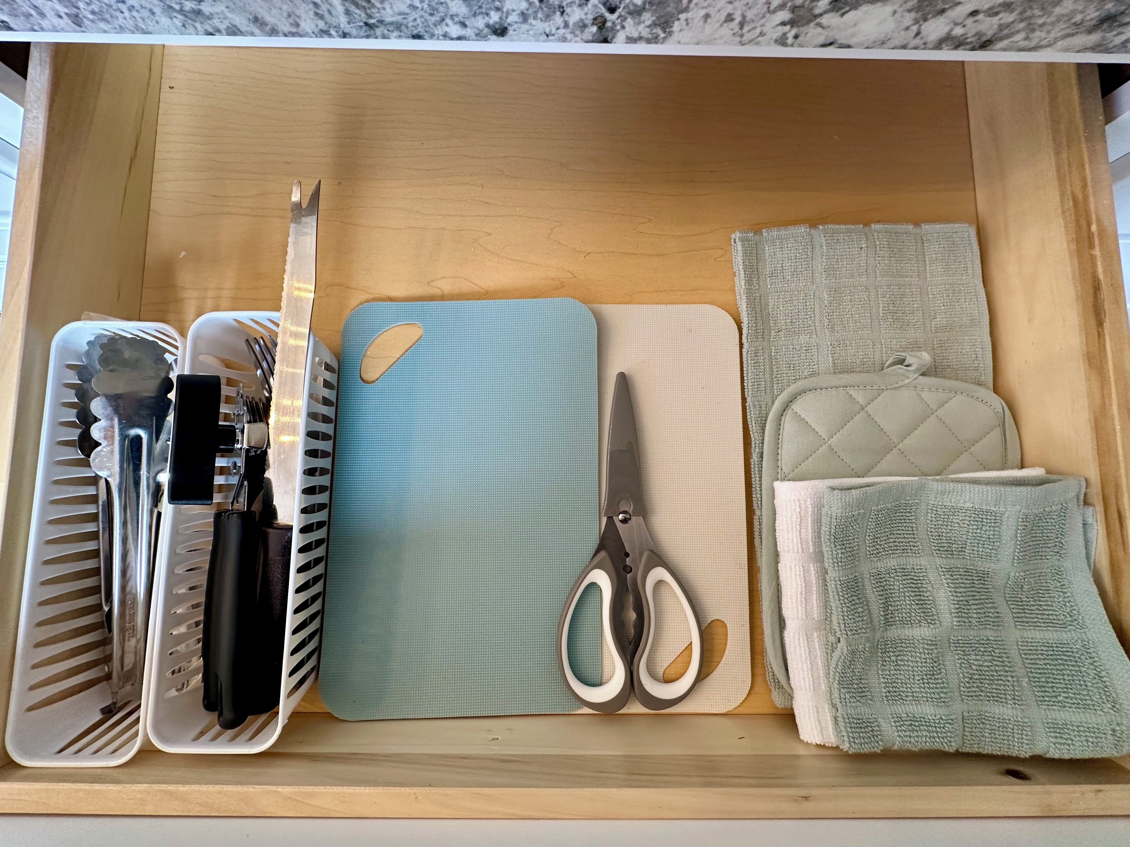 Eine Schublade mit einer blauen Unterlage, einer Schere, einem Messer mit schwarzem Griff, einer weißen Tablettgarnitur und einer grünen gesteppten Tasche.