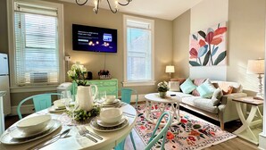 Un comedor con una mesa dispuesta para cuatro personas, un televisor montado en la pared y un sofá con un tapete floral.