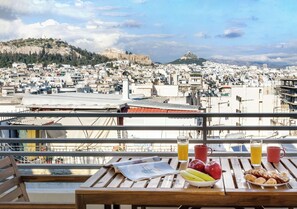 A table with breakfast items and a newspaper on a balcony overlooking a cityscape with historical buildings. - Urban Oasis with Acropolis Views by GHH (Kallithea)