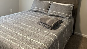 Een net opgemaakt bed met een grijs geborreld beddegoed en kussens, een houten hoofdbord, en een gevouwen grijze deken op het bed.