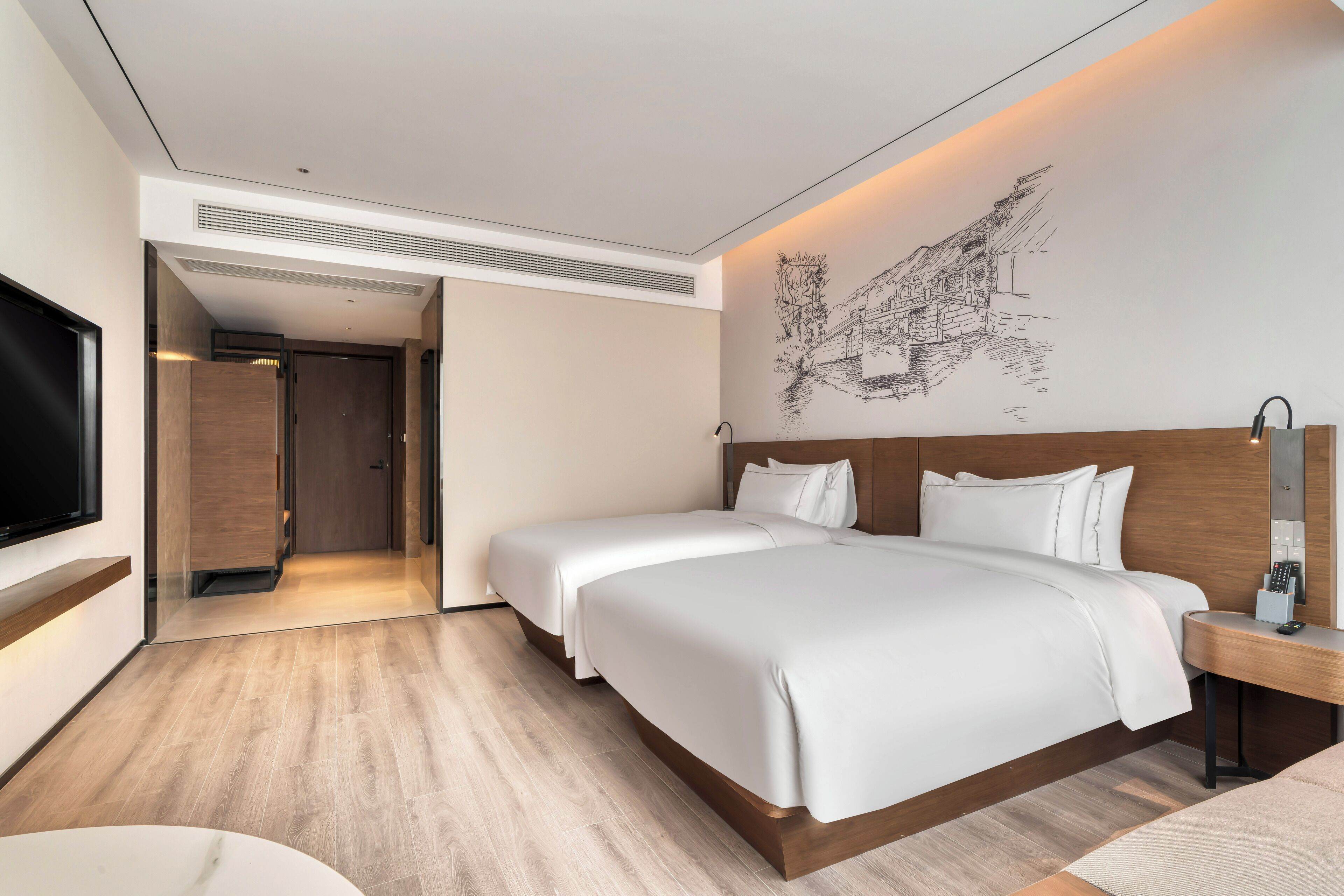 A modern hotel room with a large bed, a bedside table with a lamp, and a flat-screen TV mounted on the wall.