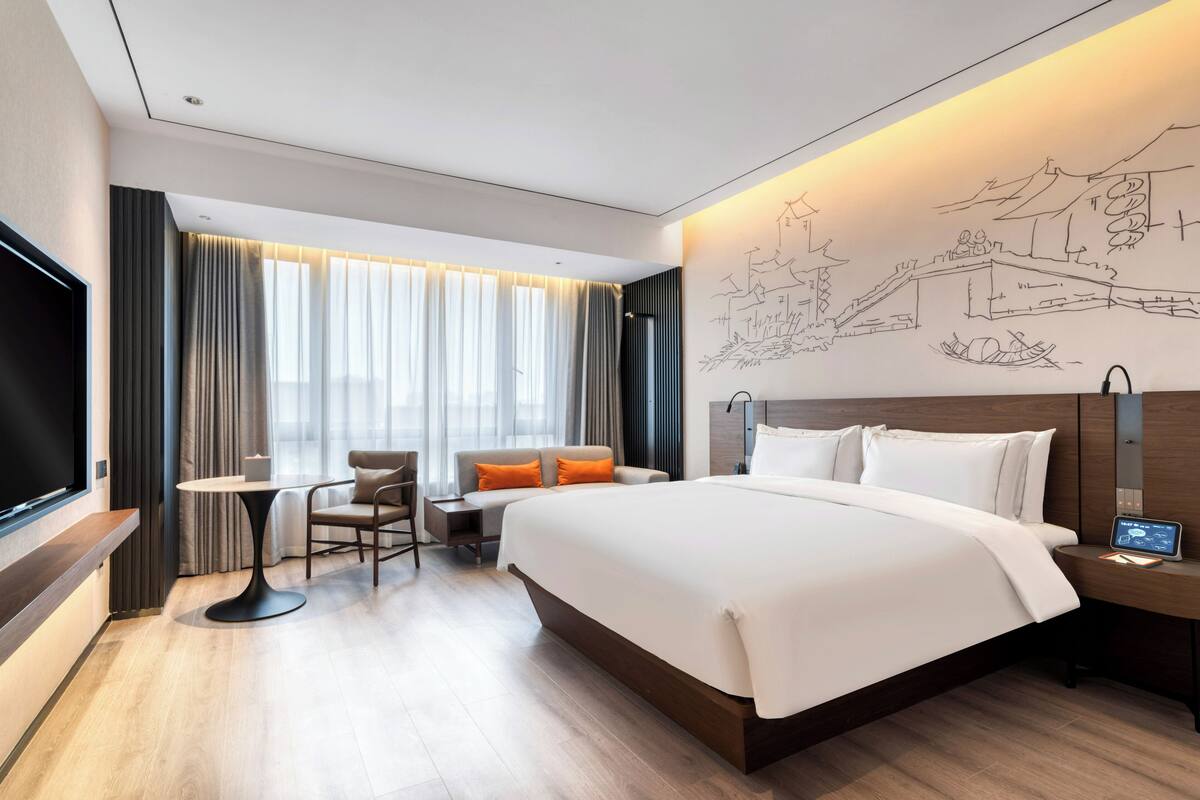 A modern hotel room with a large bed, a desk with a chair, a TV, and a wall mural.