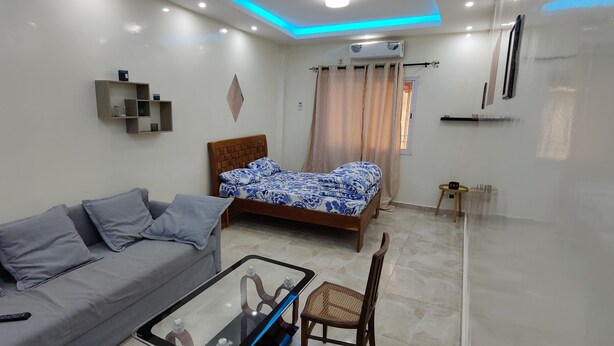 A modern living room with a grey sofa, a wooden bed with patterned bedding, a glass coffee table, and a wall-mounted TV. - OUAKAM self-contained studio 10m from the monument (Ouakam)