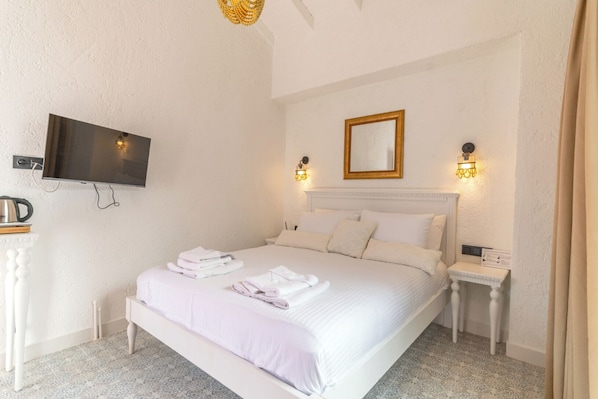 Une chambre d’hôtel bien aménagée, avec un grand lit, un téléviseur à écran plat fixé au mur, une table de chevet avec une lampe et une petite table avec une bouilloire et des livres.