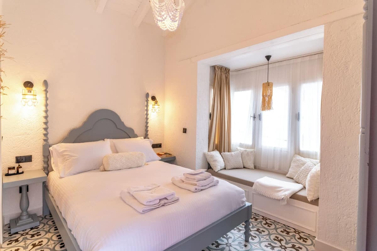 Une chambre à coucher avec un grand lit, des lampes de chevet, un coin fenêtre agrémenté de coussins et un lustre.