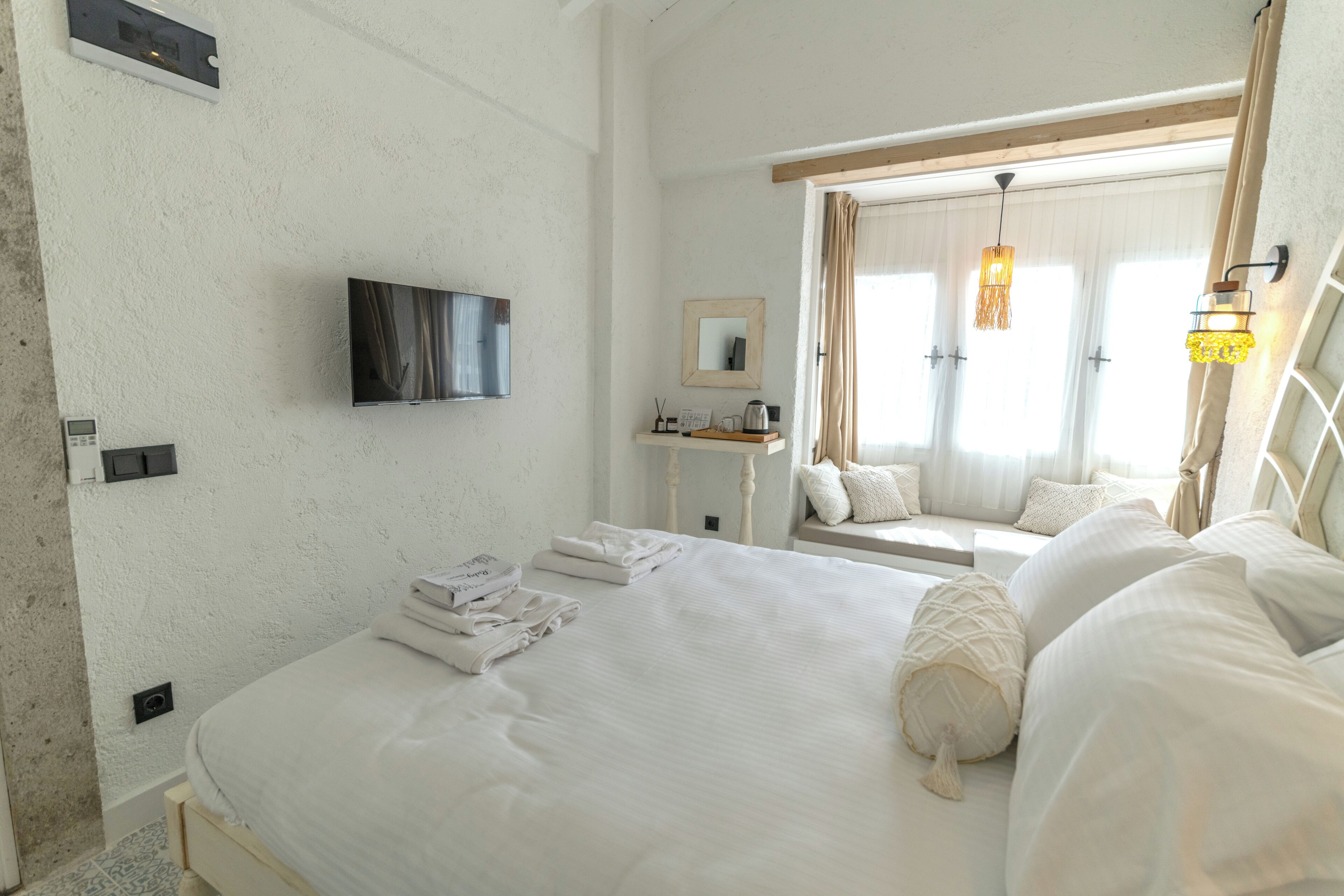 Habitación de hotel con una cama grande, un televisor instalado en la pared, una mesita de noche con lámpara y una ventana con cortinas.