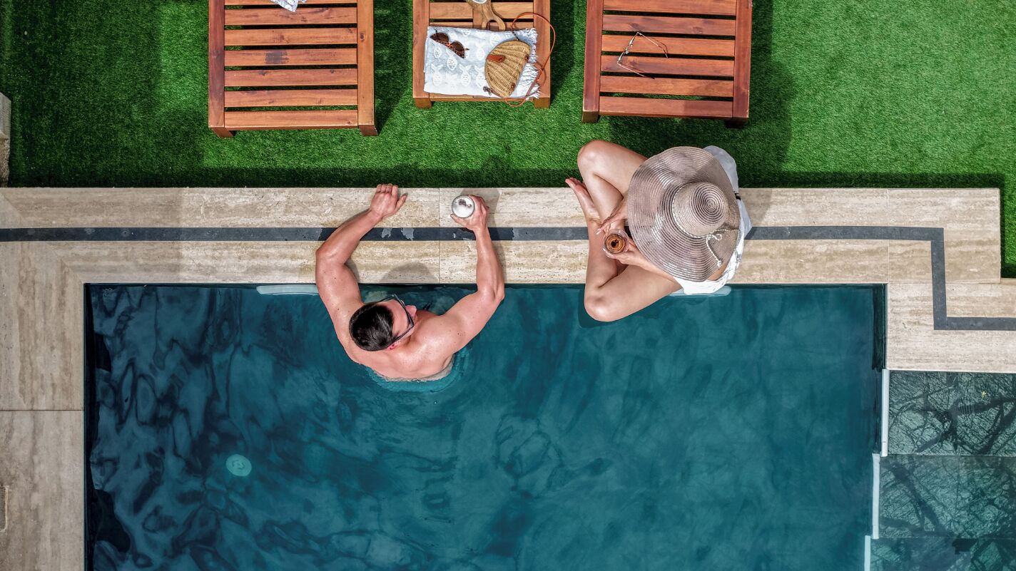 Vue plongeante d’une piscine avec deux chaises longues en bois et une table sur laquelle se trouvent un chapeau et des lunettes de soleil.