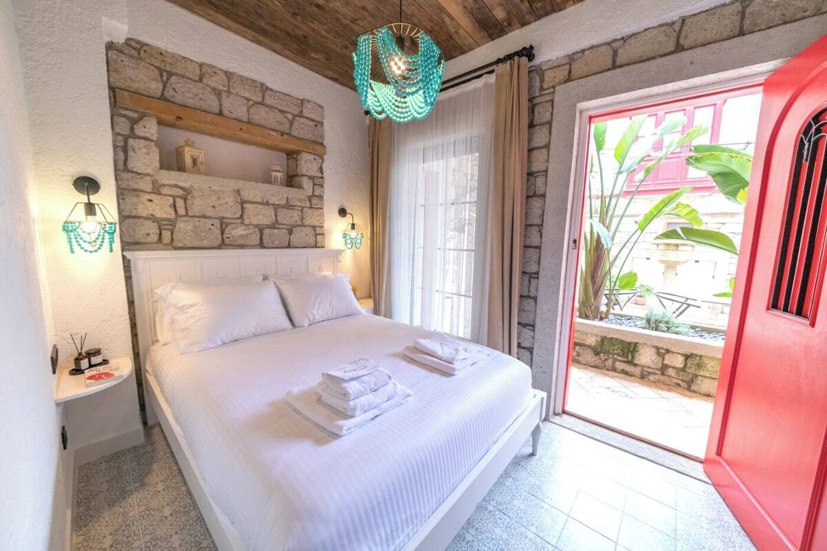Une chambre avec un mur en pierre, un lit recouvert de draps blancs, une table de chevet avec une lampe, et une vue sur l’extérieur.