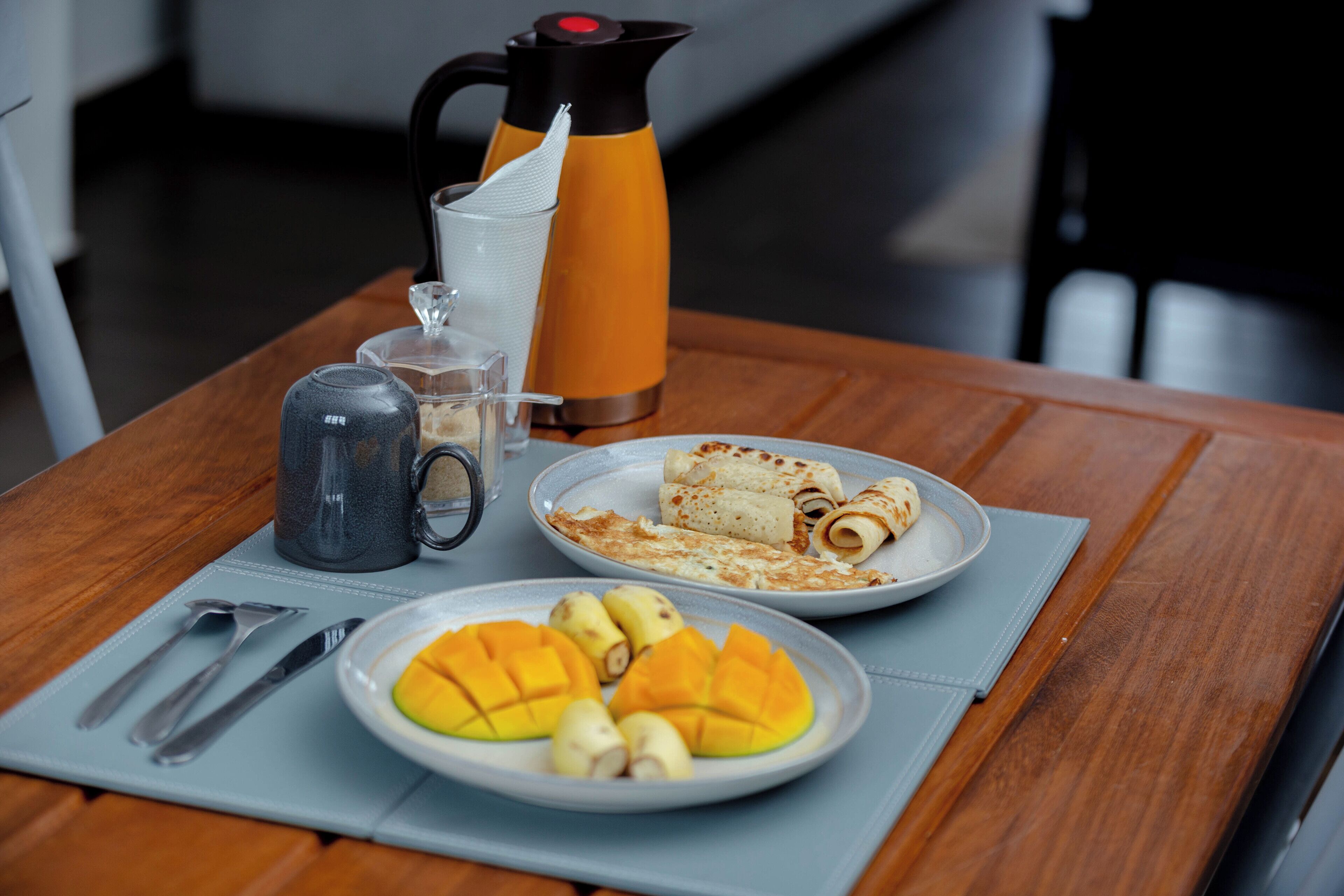 A wooden table with a coffee pot, a glass pitcher, a plate of crepes, and a plate of sliced mango.