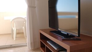 Un téléviseur posé sur un meuble en bois, avec une télécommande et un lecteur DVD, une chaise en plastique blanche et une porte-fenêtre coulissante donnant sur un balcon.