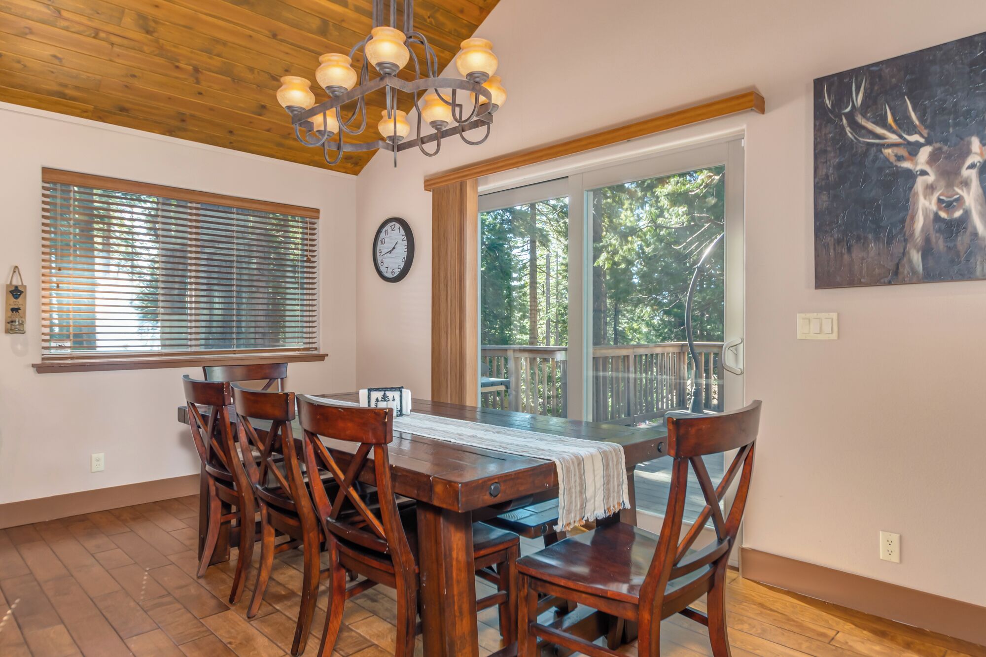 A dining area with a wooden table and chairs, a chandelier, a clock, and a large window with a view of trees.