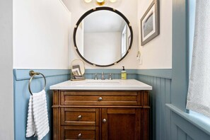 A bathroom with a wooden vanity, a round mirror, and a white sink. - Sandpiper by the Sea For 9! (Ogunquit)