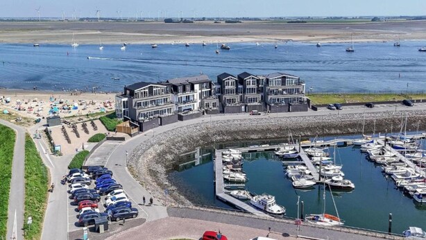 Un port de plaisance situé sur la côte, où sont amarrés de nombreux bateaux, un parking avec des voitures et un bâtiment doté de balcons donnant sur l’eau.