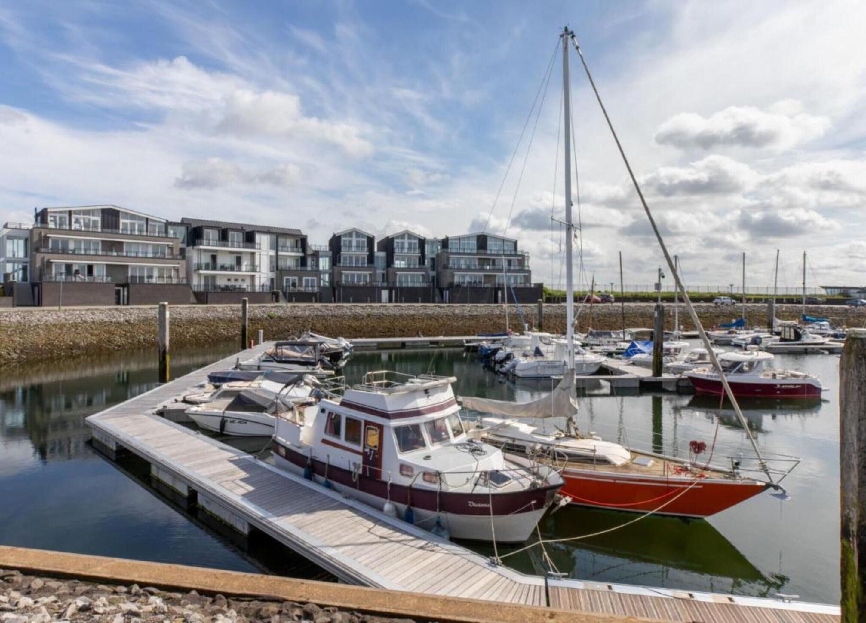 Un puerto deportivo con varias embarcaciones amarradas, un muelle de madera y edificios residenciales modernos al fondo.