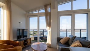 Un salon moderne avec un canapé, un fauteuil et une petite table. De grandes fenêtres offrent une vue sur la mer.