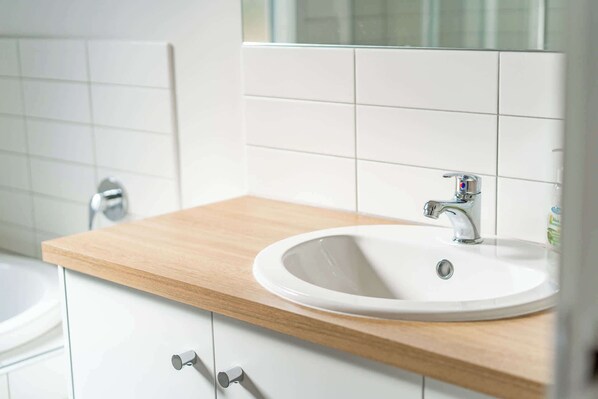 A bathroom with a white sink, a wooden countertop, a mirror, and a white tiled wall.