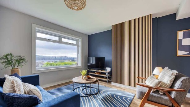 A modern living room with a large window, a flat-screen TV, a blue sofa, a wooden chair, and a striped rug.