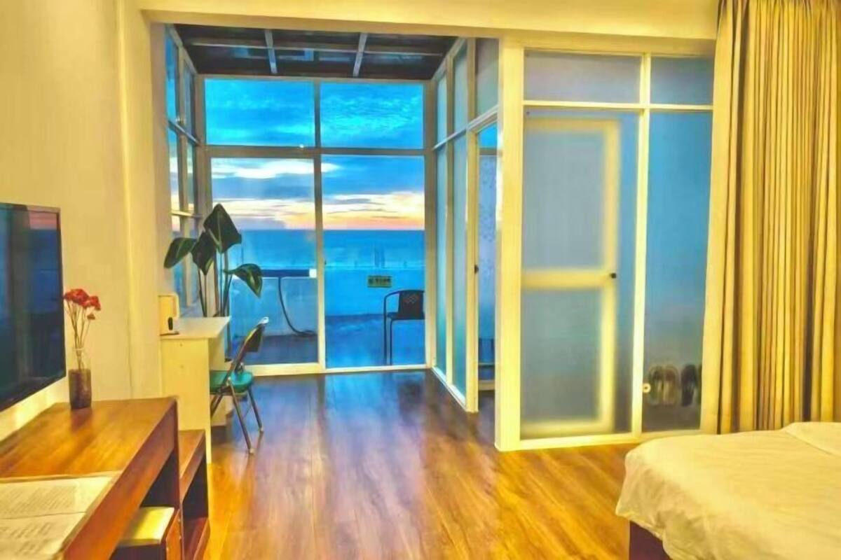 Ein Zimmer mit Holzboden, einem Bett, einem Fernseher, einem Schreibtisch, einem Stuhl, einer Pflanze und einer Schiebetür zu einem Balkon mit Meerblick.