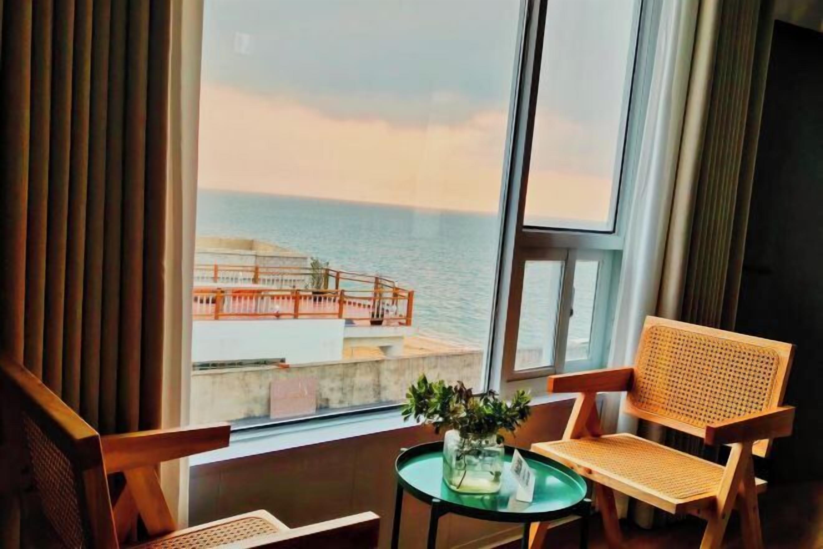 Ett rum med ett stort fönster, en trästol, ett litet bord med en vas och utsikt över havet och en avlägsen byggnad.