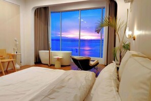 A hotel room with a large bed, a sitting area with a chair, a small table, a bathtub, and a large window with a view of the sea.