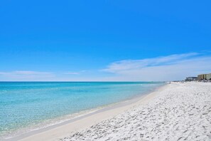 Ein Strand mit klarem türkisblauem Wasser, weißem Sand und einem Hotelgebäude auf der rechten Seite.