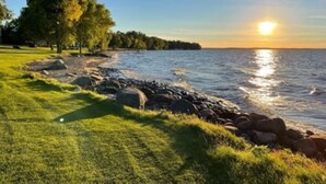 A grassy area with a rocky shoreline and trees in the background. - Relax and Recharge: Mille Lacs Lake Beach Villa - Completely Remodeled (Onamia)