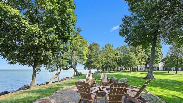 A circular outdoor seating area with Adirondack chairs and a fire pit, surrounded by a brick patio and large rocks, overlooking a body of water and a well-maintained lawn. - Relax and Recharge: Mille Lacs Lake Beach Villa - Completely Remodeled (Onamia)