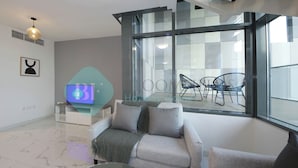 Ein modernes Wohnzimmer mit einer Couch, einem Glastisch und einem Fernseher. Ein großes Fenster bietet einen Blick auf eine Sitzgruppe im Freien.