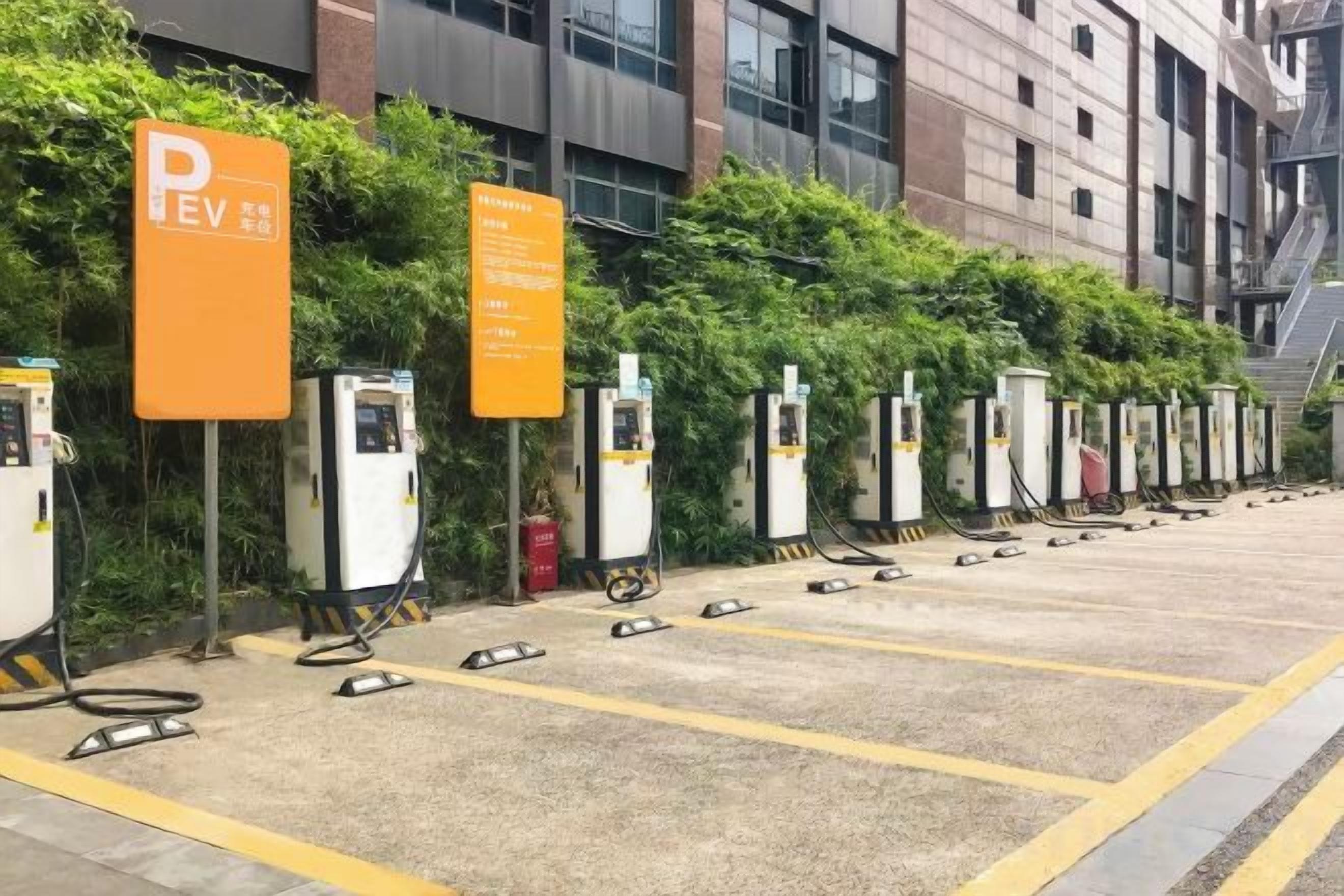 A row of electric vehicle charging stations with orange signage, set against a backdrop of a building and greenery.