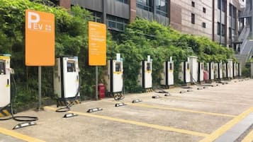 A row of electric vehicle charging stations with orange signage, set against a backdrop of a building and greenery.