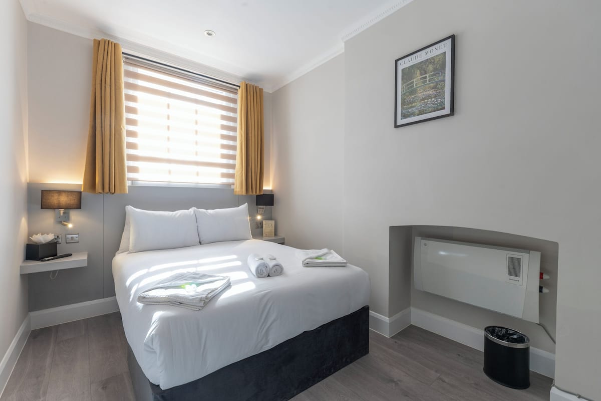 A hotel room with a bed, bedside lamps, a radiator, a framed picture on the wall, and a trash bin.