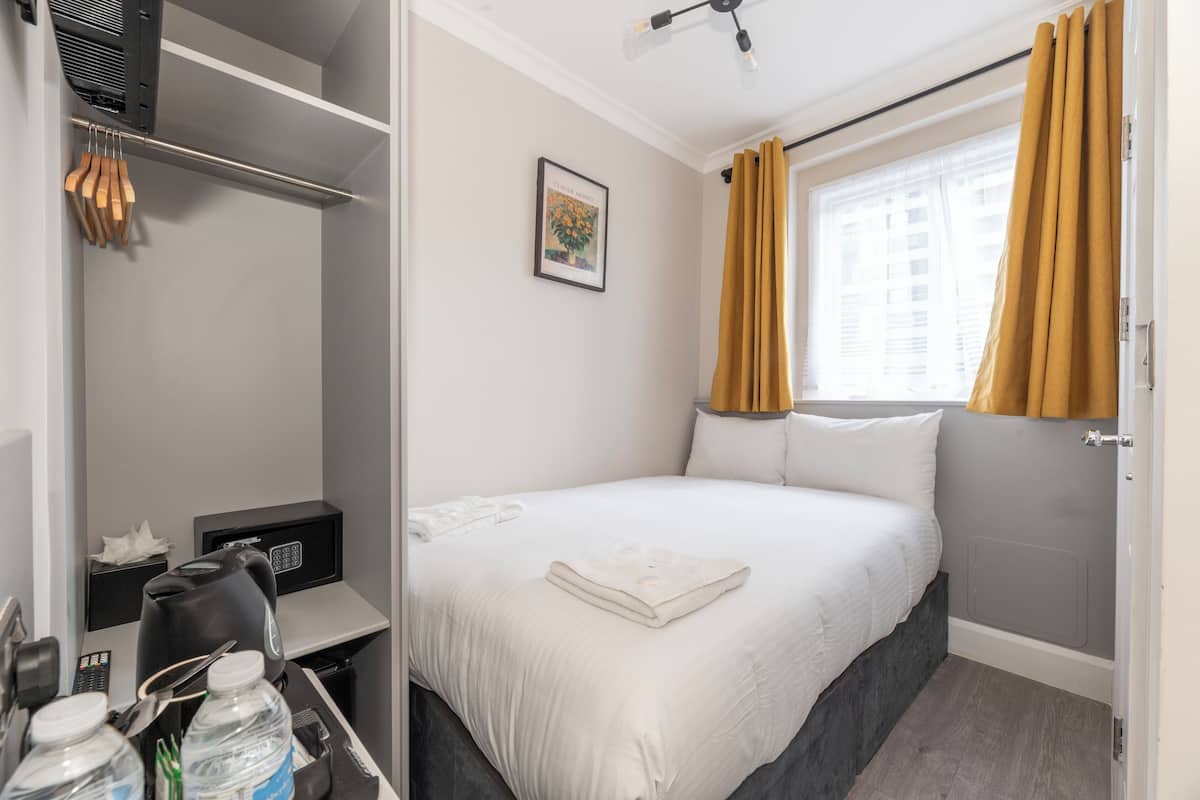 A hotel room with a bed, a desk with a kettle, a control panel, and a window with curtains.